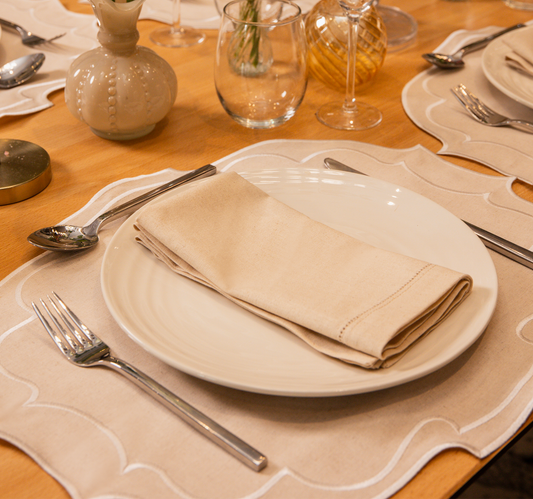 Top Tables Taylor Placemat & Napkin in beige cotton, with cream embroidered trim, styled on a wooden tables with wine and water glass with silver cutlery