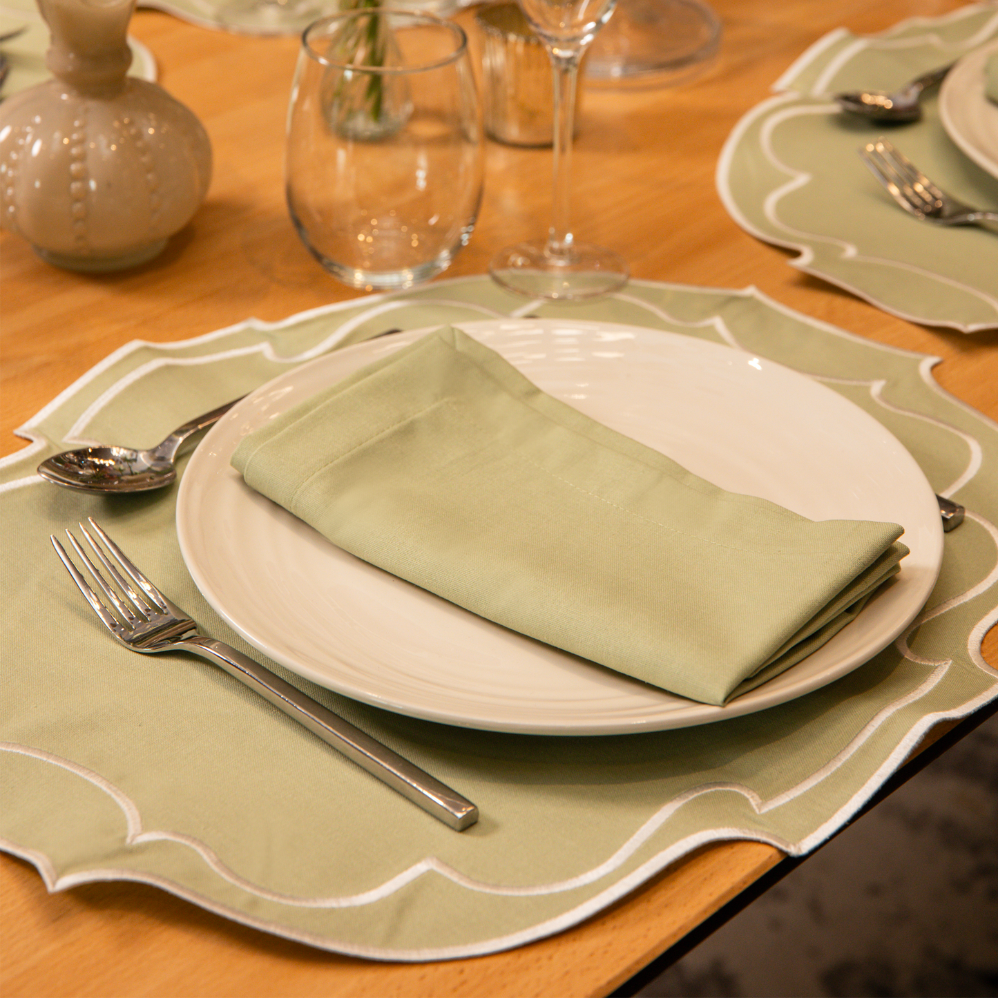 Top Tables Taylor Placemat & Napkin in Sage Green cotton, with embroidered trim, styled on a wooden table with wine and water glass.