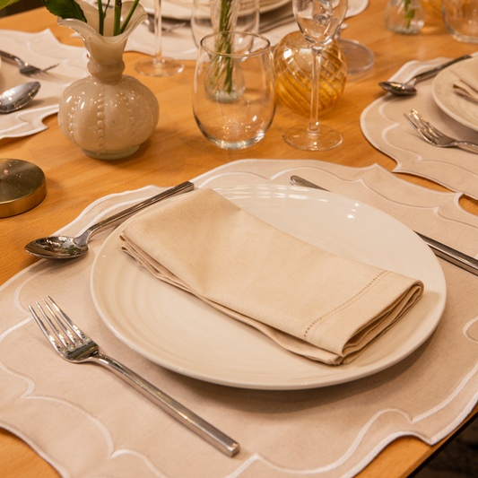 Top Tables Taylor Placemat & Napkin in beige cotton, with cream embroidered trim, styled on a wooden tables with wine and water glass with silver cutlery
