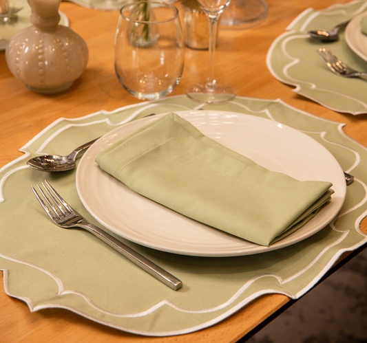 Top Tables Taylor Placemat & Napkin in Sage Green cotton, with embroidered trim, styled on a wooden table with wine and water glass. 