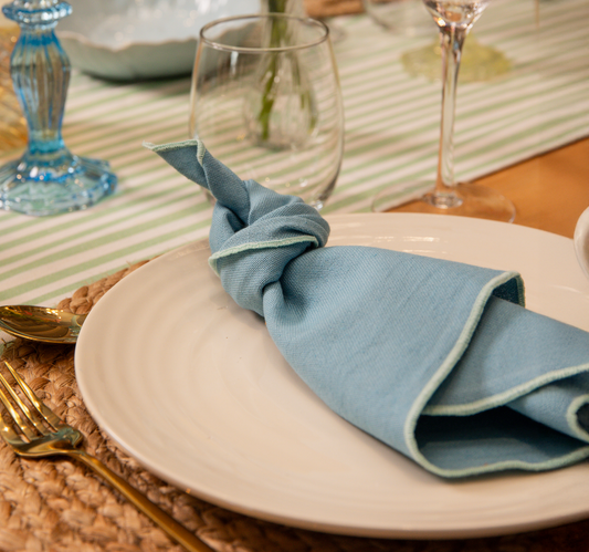 Top Table Tilly Linen cotton napkin in blue with aqua edge stitching. Placed on a Jute placemat with cream plate and gold cutlery